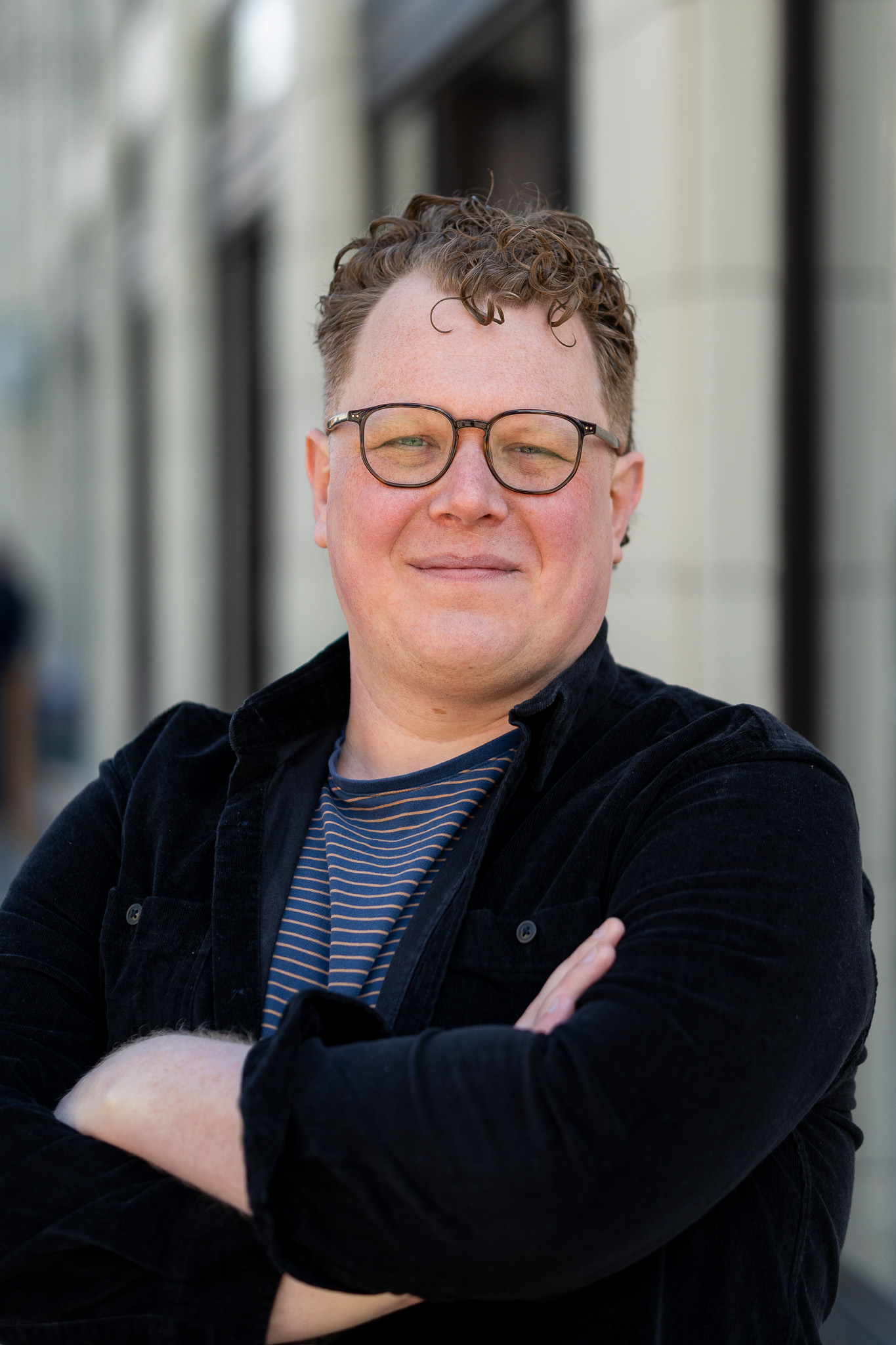 Headshot of Jake Bakker, founder of JBakker Web Dev. Jake is smiling with his arms crossed professionally. They are white with short curly hair, wearing glasses, a solid dark flannel and a blue and brown striped shirt underneath